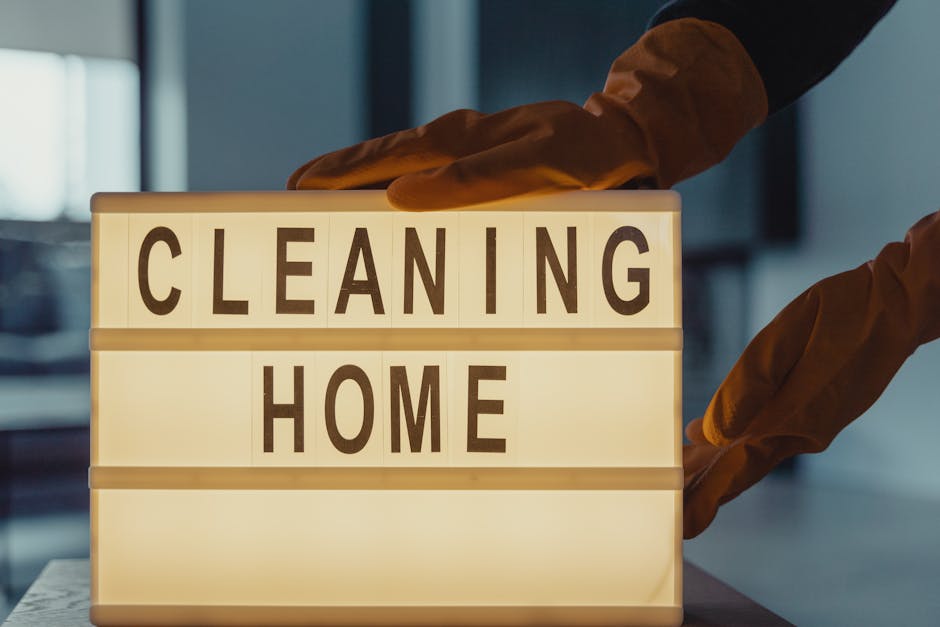 A smiling woman dressed in a bright yellow uniform with yellow gloves, holding a spray bottle in one hand and a blue cleaning cloth in the other, standing in a well-lit, modern living room. The room f